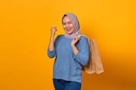 Cheerful woman in hijab holding shopping bags with a joyful expression against a vibrant yellow background.