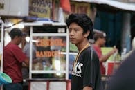 A teenage boy in a bustling street market with food vendors in the background.