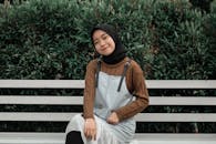 Young Asian woman in hijab sitting on a bench, smiling against lush greenery, exuding a peaceful outdoor vibe.
